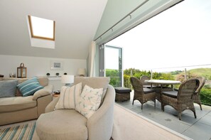 A modern living room with a sofa, a dining table with chairs, a large window, and a view of greenery.