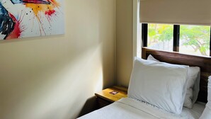 Habitación de hotel con cama, mesita de noche, ventana con vista a follaje y un cuadro abstracto colorido en la pared.