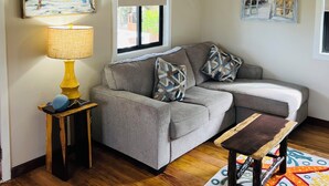 Sala de estar con sofá gris, cojines estampados, una pequeña mesa de madera con lámpara y una alfombra colorida.