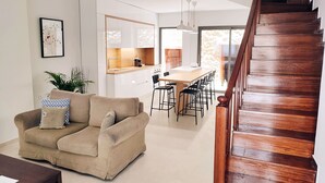 A modern living room with a sofa, a dining area with chairs, a kitchen with white cabinets, and a wooden staircase.