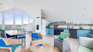 A spacious living area with wicker furniture, a dining table, and a kitchen with blue cabinets and a large window overlooking the countryside. - The Point (Wadebridge)