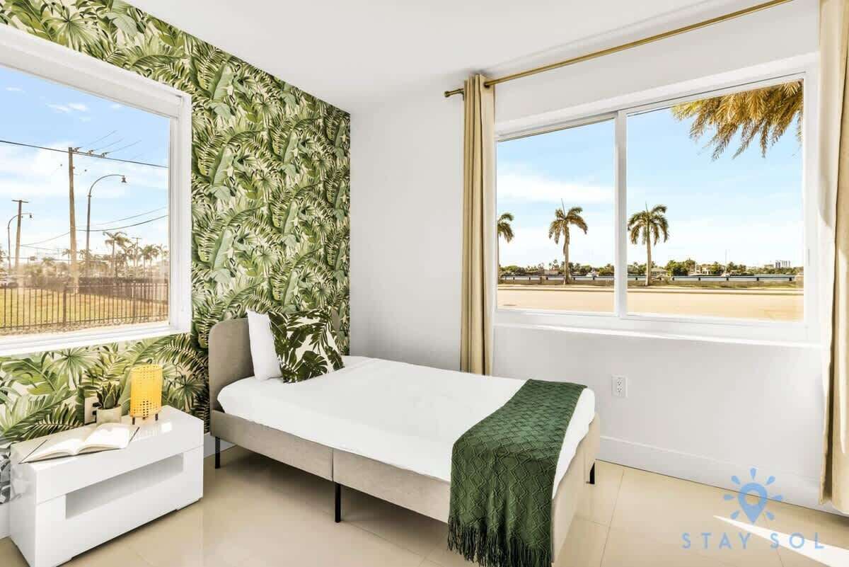A room with a bed, a window with a view of palm trees, and a tropical wallpaper.