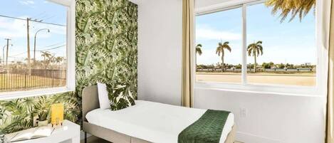 A room with a bed, a window with a view of palm trees, and a tropical wallpaper.