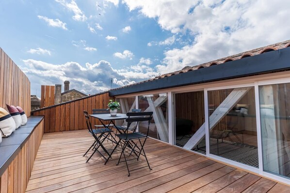 A wooden deck with a table and chairs, a potted plant, and a sliding glass door leading to an interior space. - Volubilis Guest House (Bordeaux)