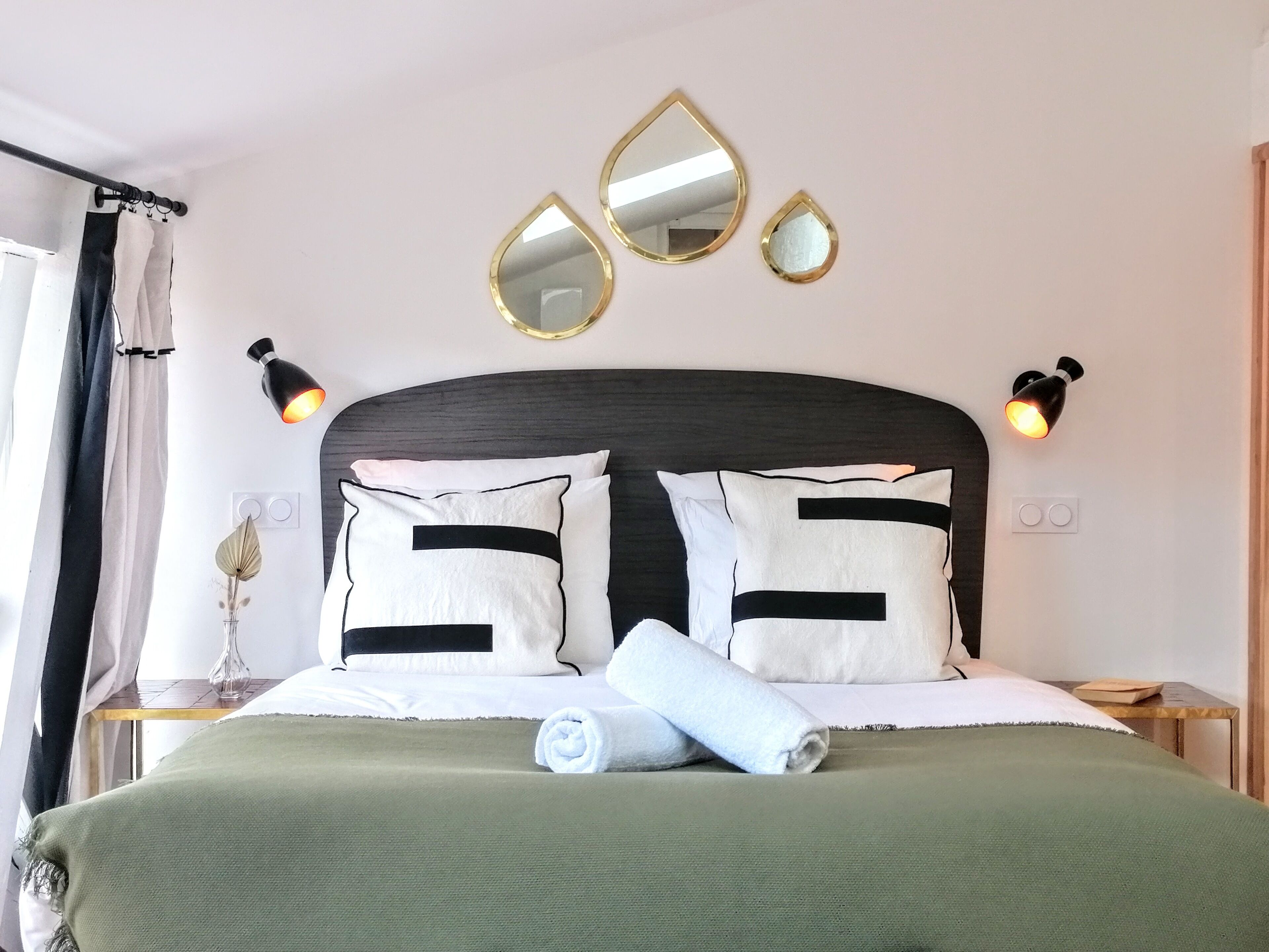 A neatly made bed with white and black pillows, a green blanket, and a rolled towel. There are two wall-mounted mirrors and two wall sconces.