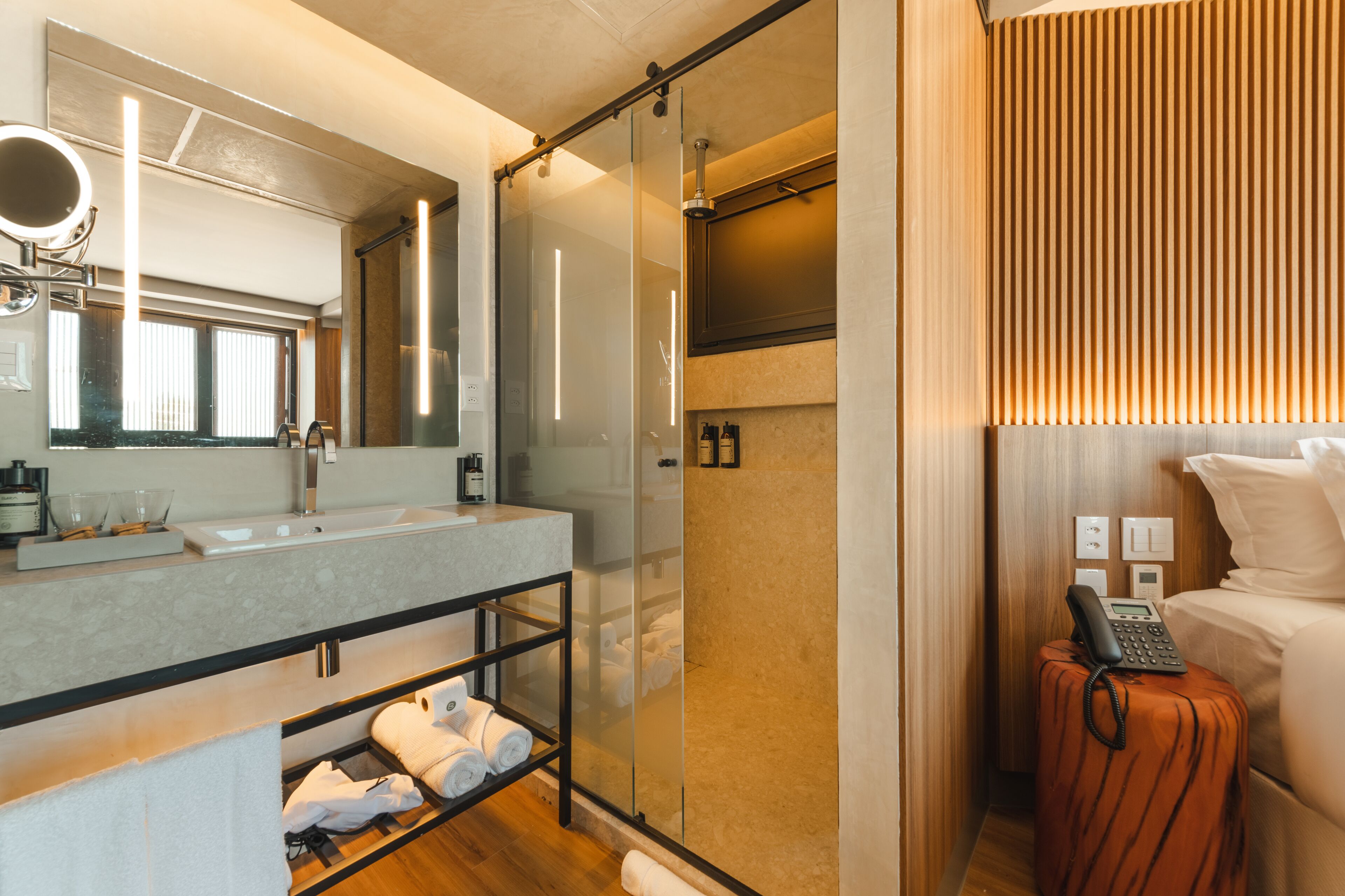 A modern bathroom with a glass shower enclosure, a vanity with a mirror, and a bedside table with a phone.