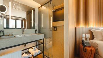 A modern bathroom with a glass shower enclosure, a vanity with a mirror, and a bedside table with a phone.