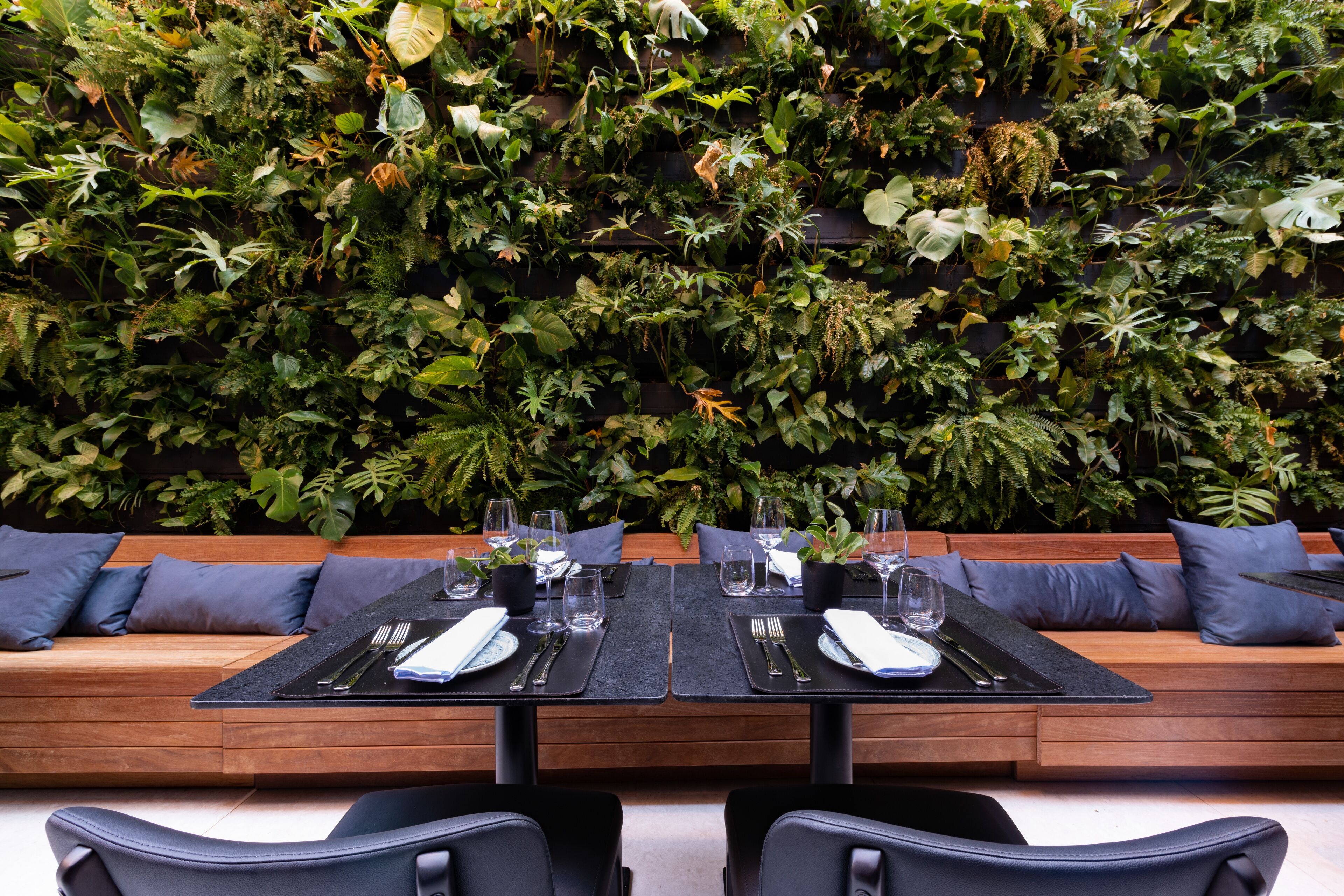 A dining area with a long wooden bench and two tables set for two, each with a black placemat, cutlery, and glasses.