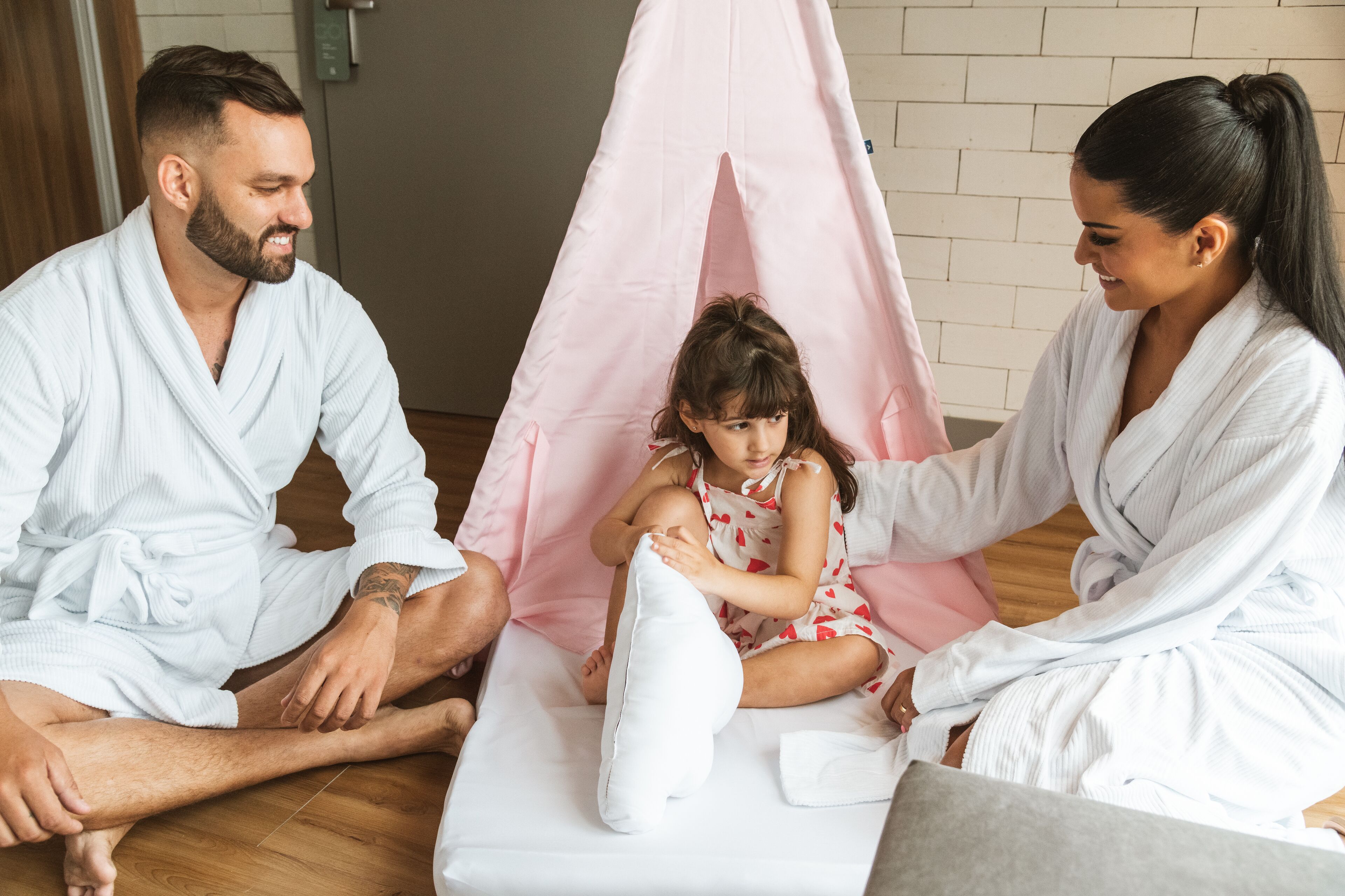 A family of three in a room with a wooden floor and a white brick wall.