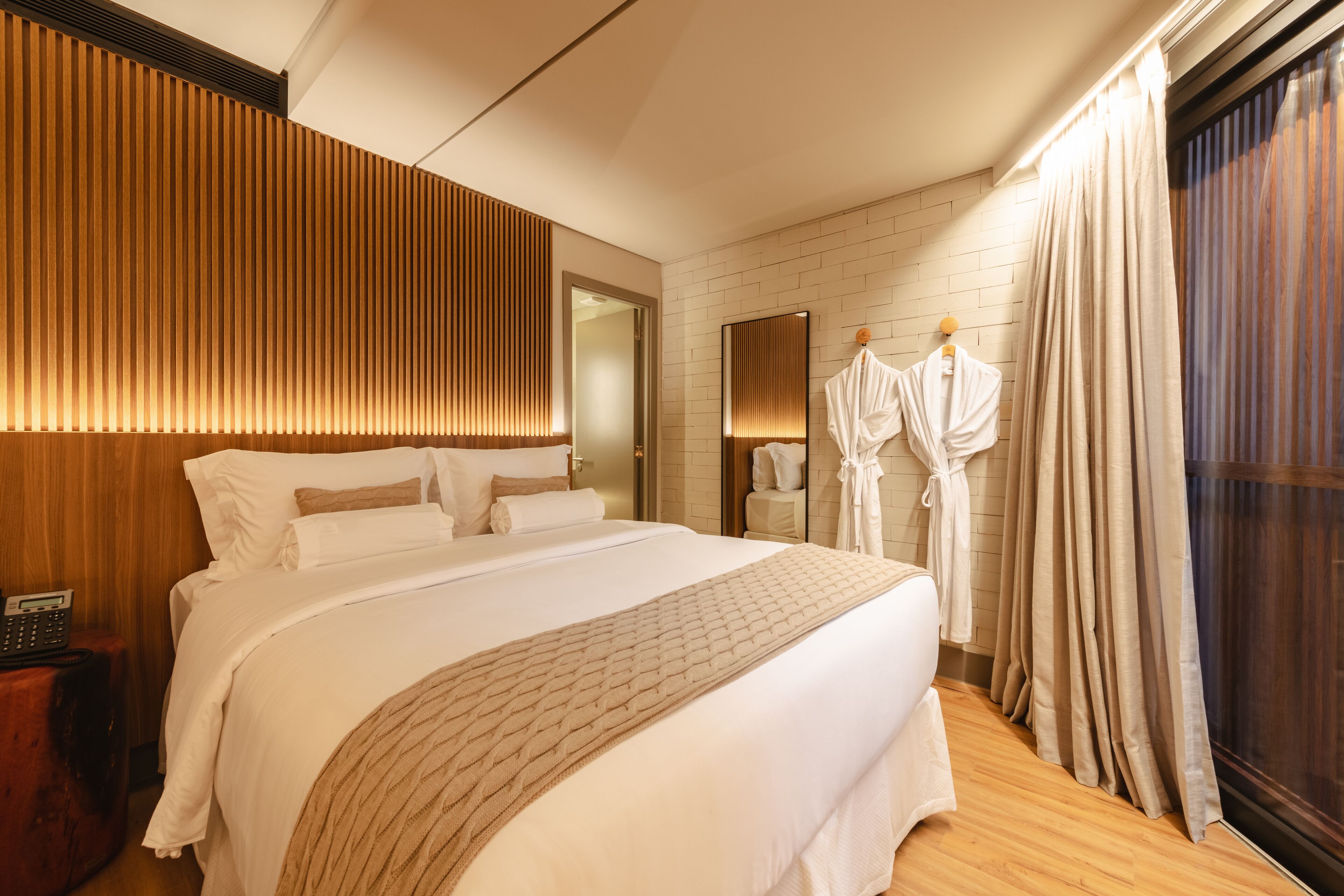 Una habitación de hotel con una cama grande, cabecera de madera, un espejo, bata y ventana con cortinas.