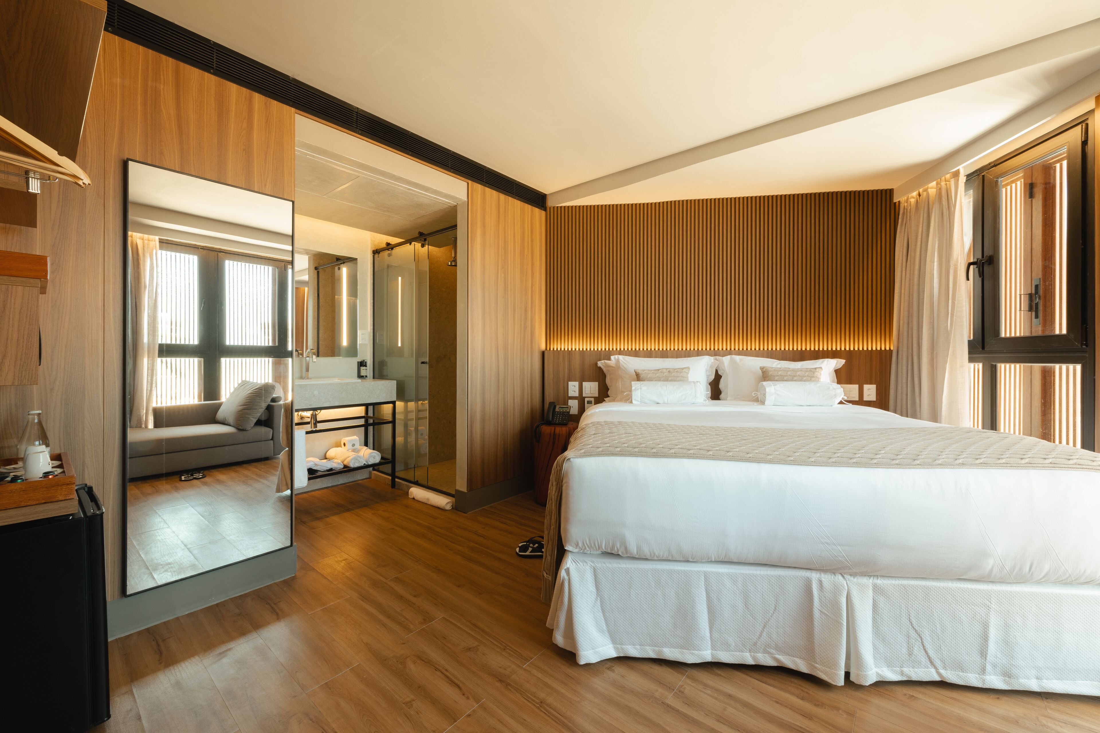 A modern hotel room with a large bed, a sitting area with a sofa and table, a large mirror, and wooden paneling.