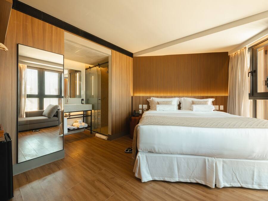 A modern hotel room with a large bed, a sitting area with a sofa and table, a large mirror, and wooden paneling.