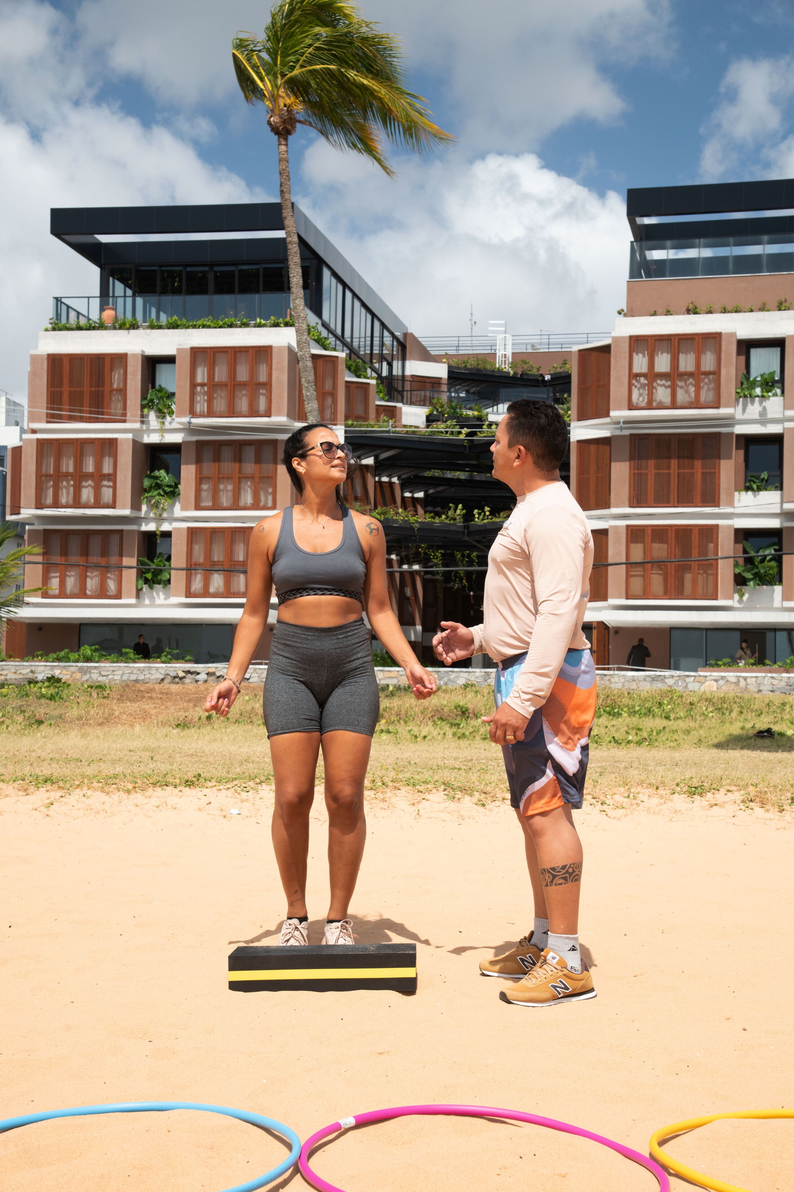 Twee personen staan op het strand, met een modern gebouw op de achtergrond.