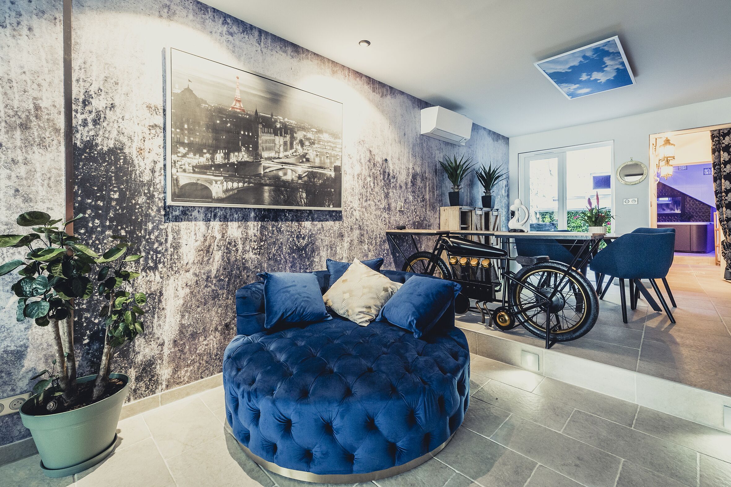 Ein modernes Wohnzimmer mit einem blauen, gesteppten Sofa, einem Fahrrad und einem Essbereich mit Tisch und Stühlen.