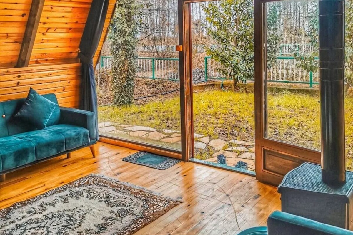 A cozy cabin interior with a wooden ceiling, a blue sofa, a stone fireplace, and a large glass door leading to a garden.