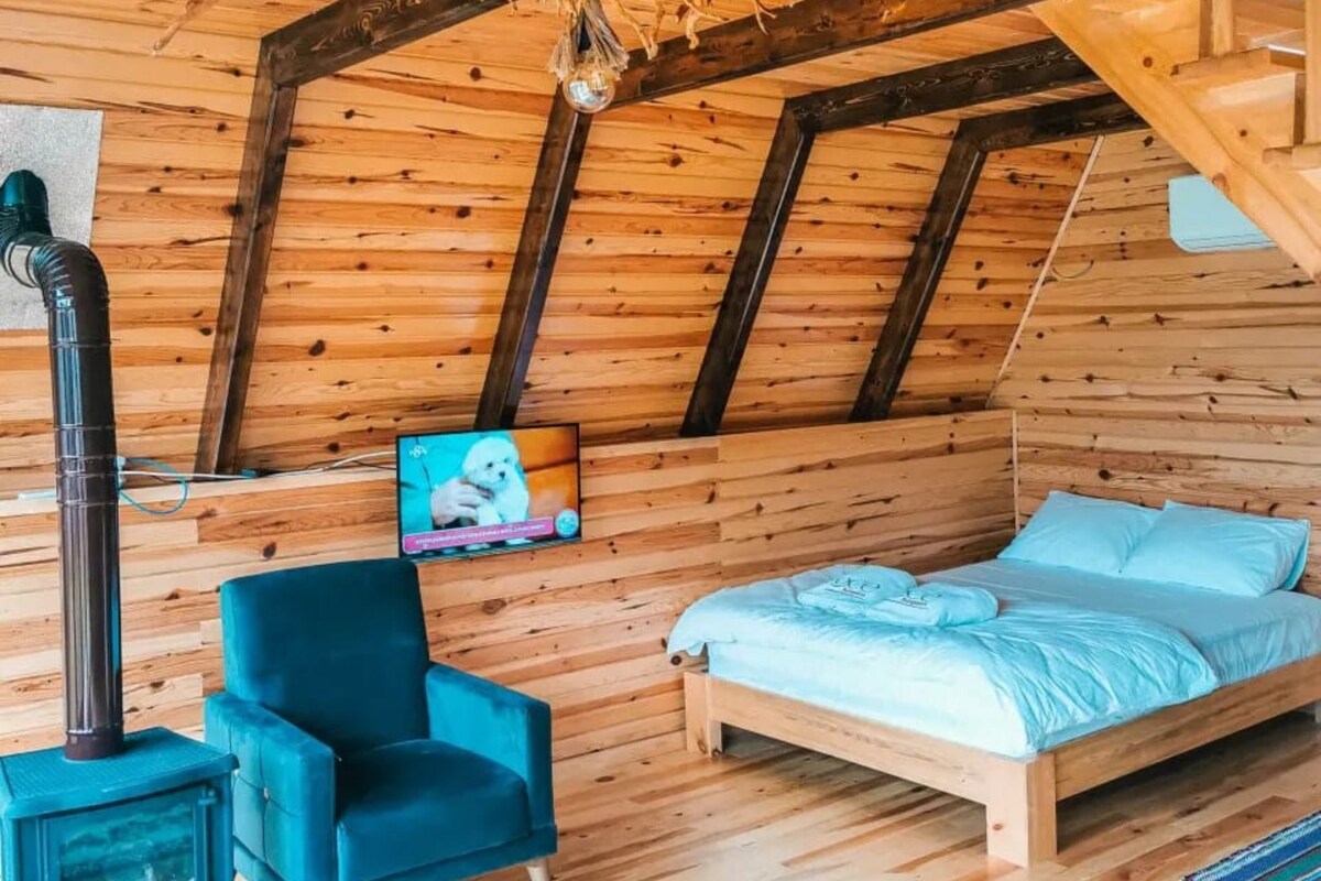Un dormitorio acogedor en el ático con techo inclinado, paredes de madera, una cama, un sillón azul y un pequeño calentador.