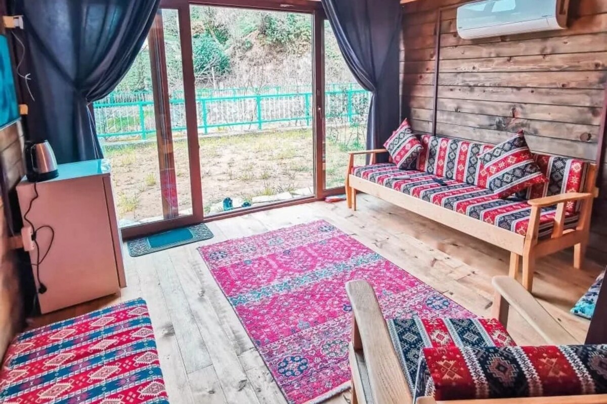 A cozy room with a wooden bench, a sofa with patterned cushions, a large rug, and a view of a green fence outside.