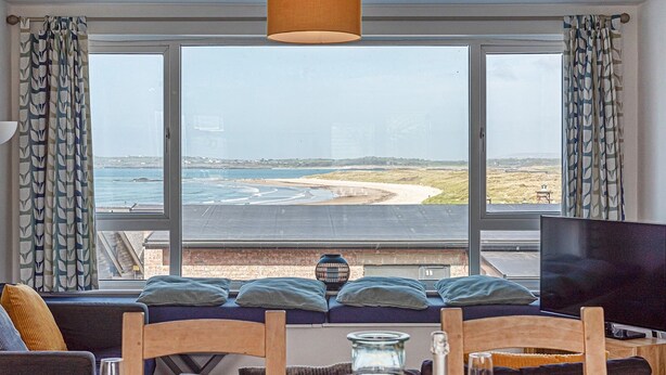 Une pièce dotée d’une grande fenêtre donnant sur une plage et la mer, d’un canapé avec des coussins, d’une table à manger avec des chaises, d’un téléviseur et d’une bouteille de vin.