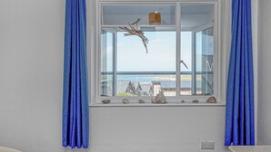 Une chambre dotée d’une grande fenêtre donnant sur la mer, comprenant un lit, une petite table et une étagère ornée d’objets décoratifs.