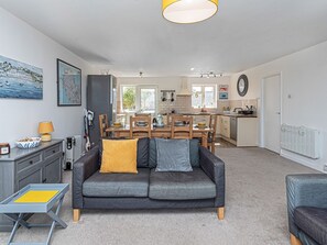 A modern living room with a grey sofa, a dining table, and a kitchen in the background. - Sea View Apartment in centre of Rhosneigr - Pass the Keys (Rhosneigr)