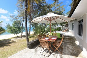 Eine Terrasse mit Tisch und Stühlen, einem weißen Sonnenschirm und Blick auf Strand und Meer.