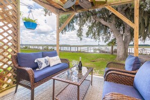 A covered outdoor seating area with wicker furniture and a glass table, overlooking a body of water.