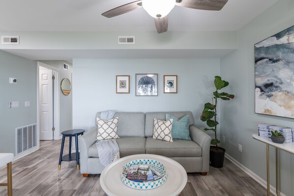 A modern living room with a grey sofa, a round coffee table, and a ceiling fan. There are three framed pictures on the wall, a potted plant, and a small table with a vase.