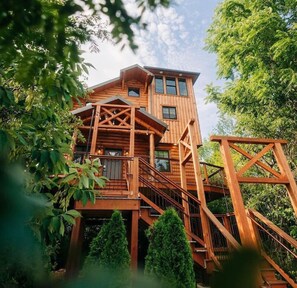 A multi-level wooden cabin with a staircase leading to the upper floor, surrounded by trees. - 3-Story Luxury Treehouse with Hot Tub - Sleeps 6 (Millersburg)