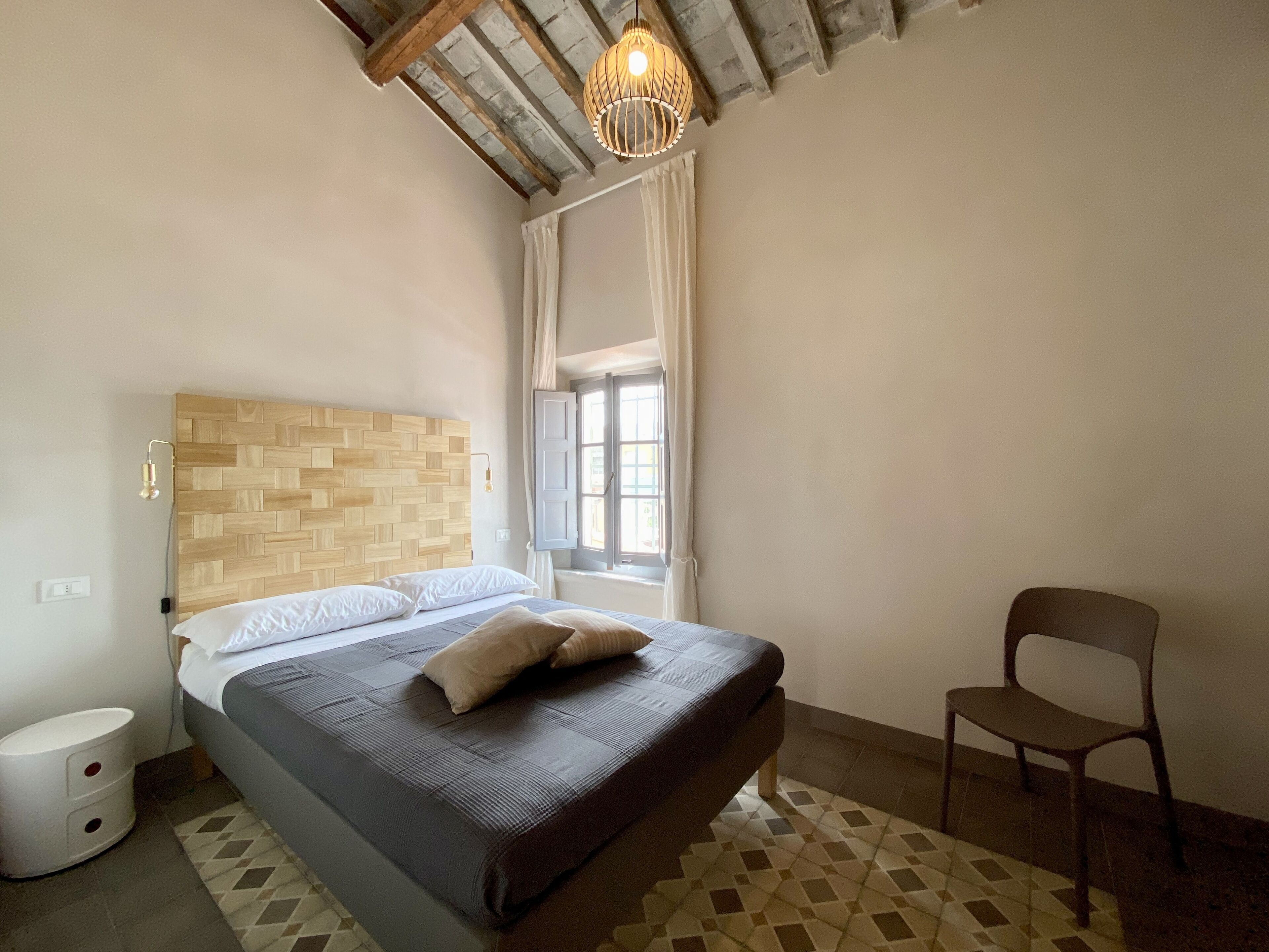 A bedroom with a wooden headboard, a bed with a dark cover, a chair, a window with curtains, and a hanging light fixture.