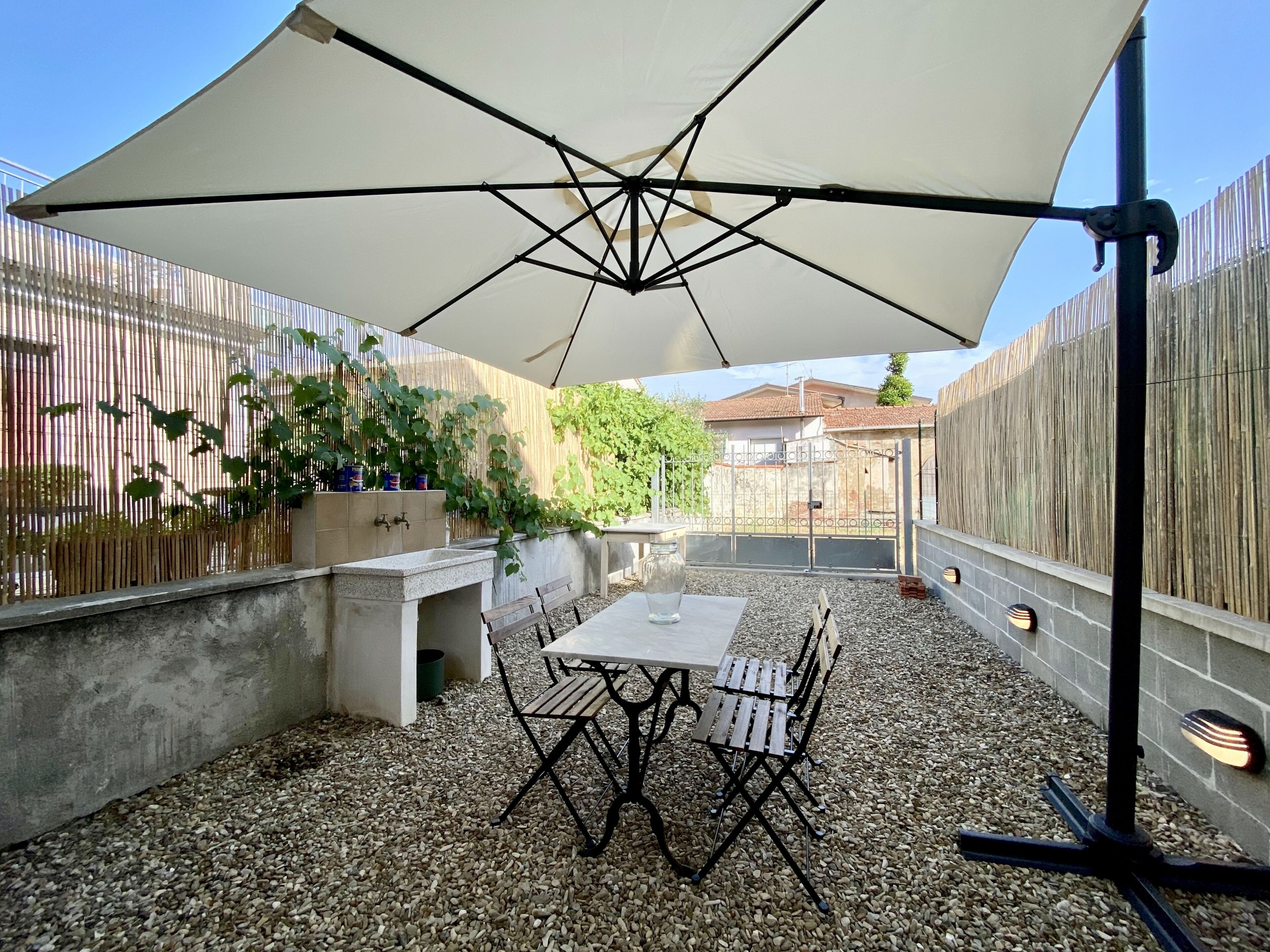Eine Terrassenanlage im Freien mit Tisch und Stühlen unter einem großen Sonnenschirm, umgeben von einem Holzzaun und einer Gartenmauer.