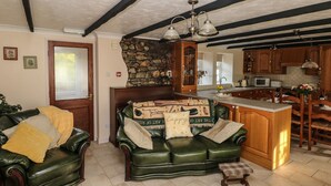 A cozy living room with a green leather sofa, a small wooden stool, and a patterned rug. A stone fireplace with a wooden mantel is visible, and the room has wooden beams on the ceiling. - Y Cartws (Llandysul)