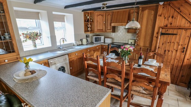A kitchen with wooden cabinets, a granite countertop, a dining table with chairs, and a window with a flower arrangement. - Y Cartws (Llandysul)