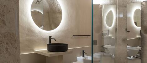 Un baño moderno con un espejo redondo, un lavabo negro y una cabina de ducha de vidrio.