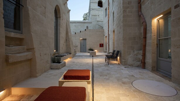 Un patio estrecho con muros de piedra, un área de estar central con bancos y una puerta que da a otro espacio.