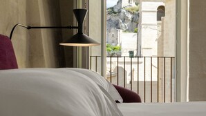 Habitación de hotel con cama, mesita de noche y ventana con vistas a una montaña rocosa y un edificio.