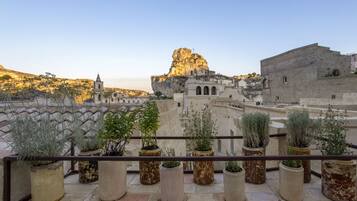 A balcony with potted plants, a view of a historic building with a prominent rock formation, and a clear sky.