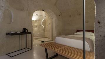 Una habitación moderna con cama, un banco de madera, una mesita pequeña y cabina de ducha con mampara de vidrio.