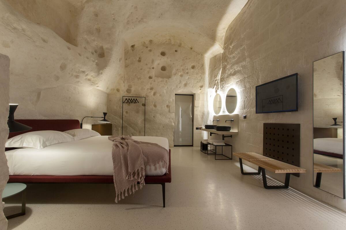 Ein modernes Hotelzimmer mit einem großen Bett, einer Bank, einem Schminktisch mit Spiegel und einem an der Wand befestigten Waschbecken.