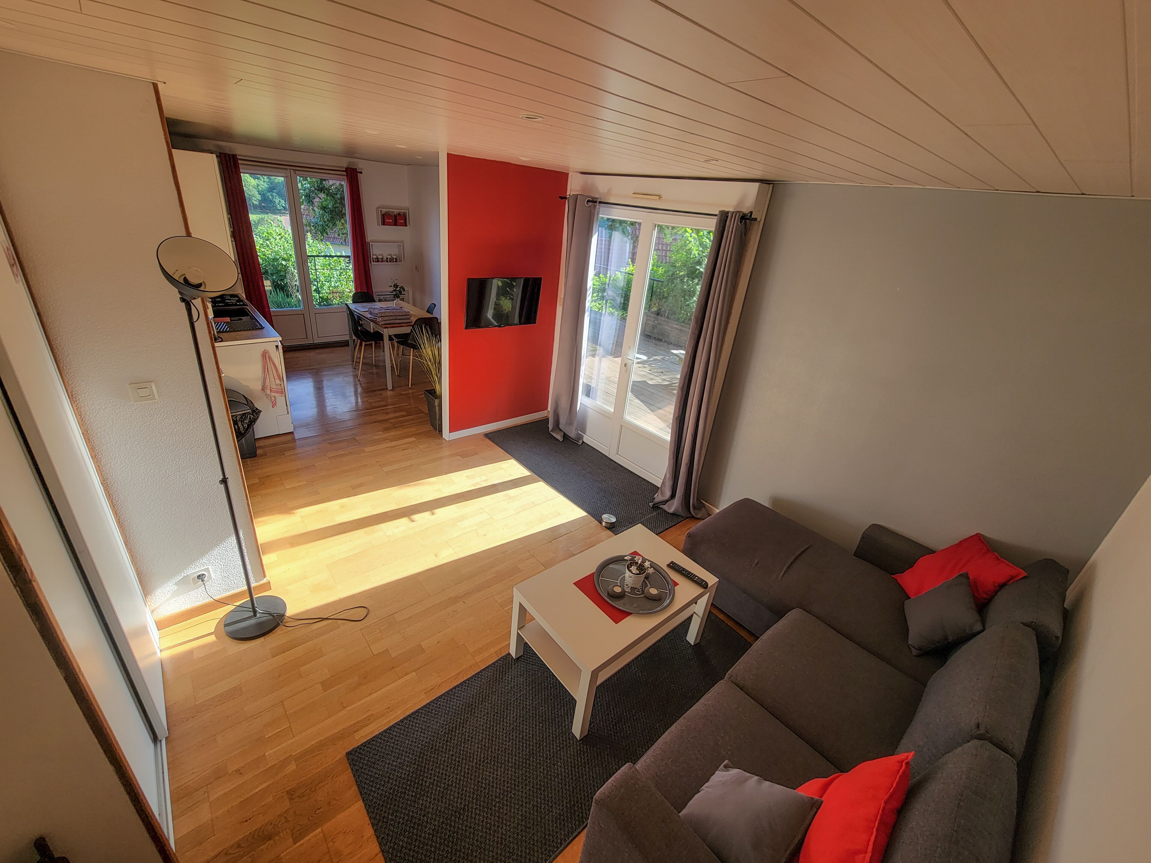 Ein modernes Wohnzimmer mit grauem Sofa, einem kleinen weißen Tisch und einem Fernseher. Durch eine offene Tür ist ein Essbereich mit Tisch und Stühlen zu sehen.