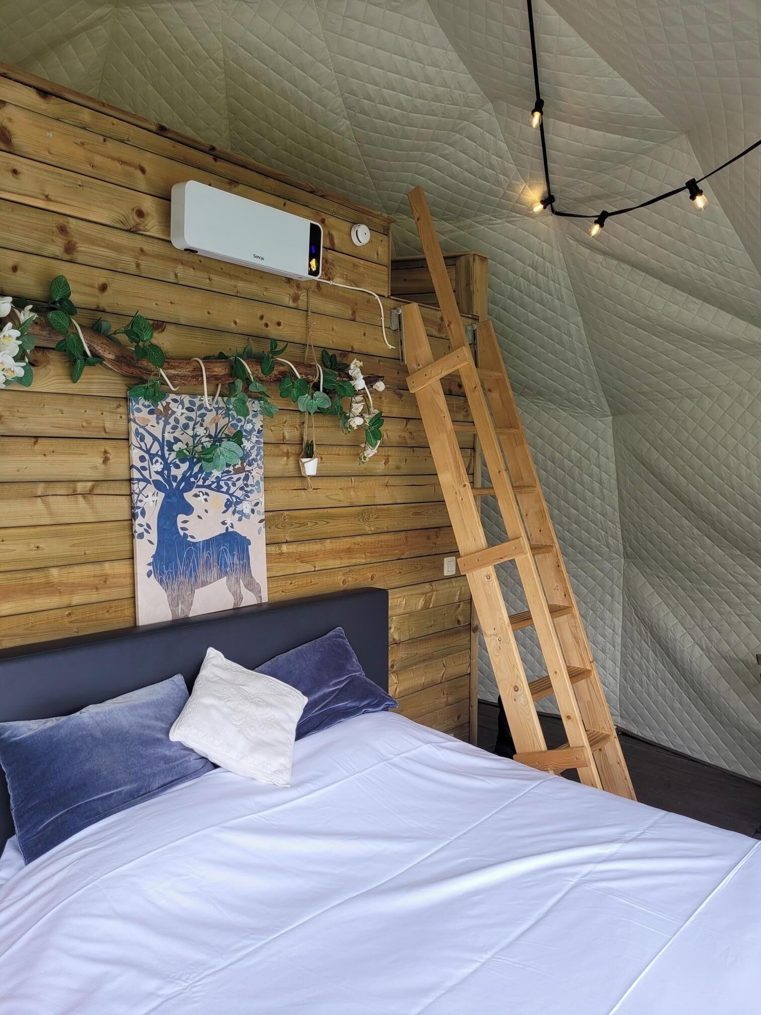 Ein Schlafzimmer mit einer Wand aus Holzlattung, einem Bett mit weißer und blauer Bettwäsche, einer Leiter und Lichterketten.