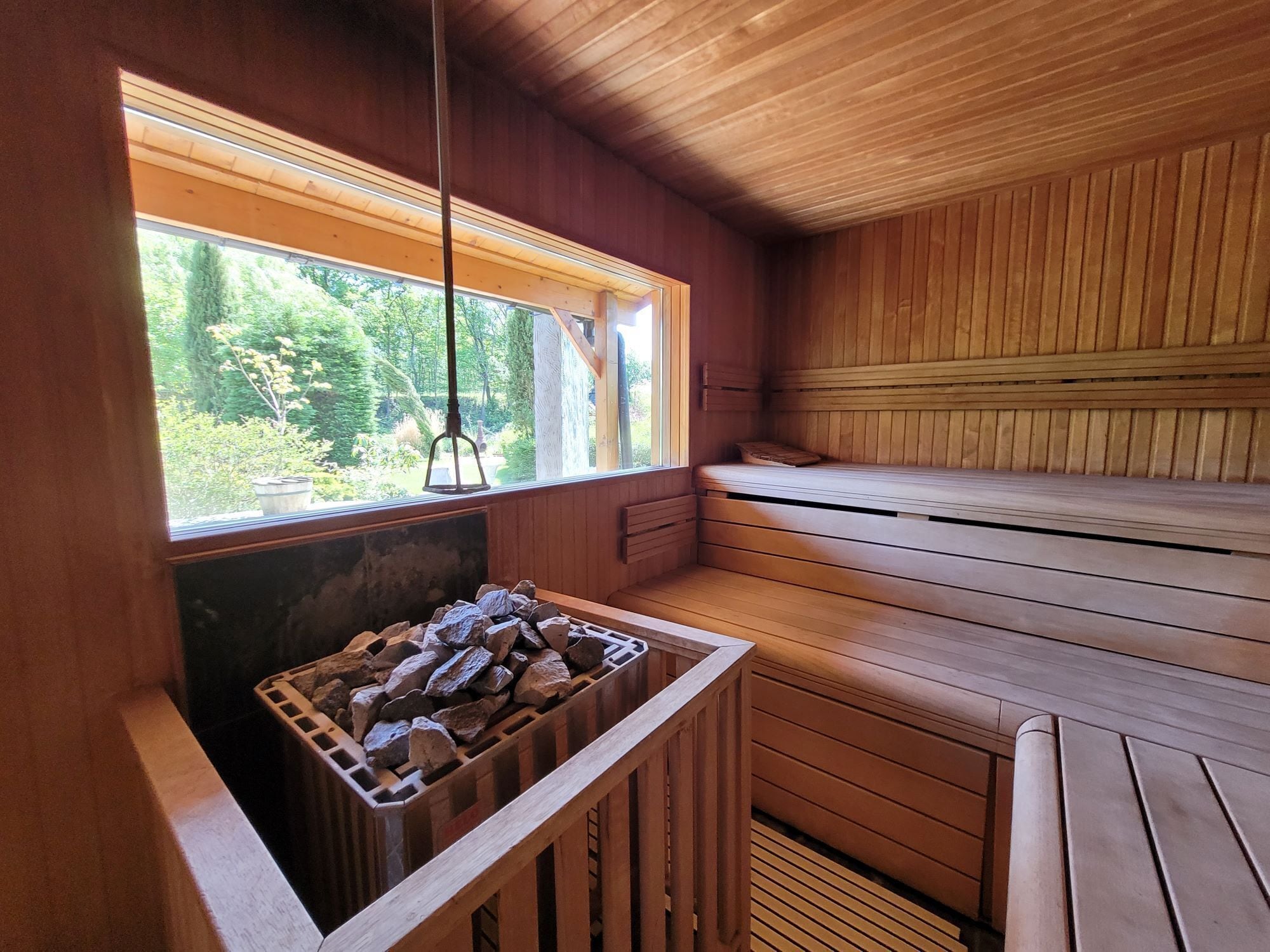 Una sala de sauna con bancos de madera, una ventana con vistas a follaje y un banco de madera con una cesta de rocas.