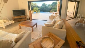 Uma sala de estar ampla com um sofá grande, uma mesa de centro e uma televisão. Há vista para um jardim através de uma porta de vidro.