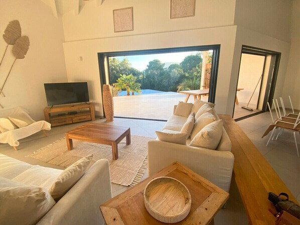 A spacious living room with a large sofa, a coffee table, and a TV. There is a view of a garden through a glass door.