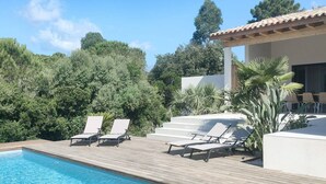 Piscina com deck de madeira e espreguiçadeiras, cercada por vegetação e um edifício com telhado telhado.