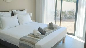 Quarto de hotel com uma cama grande, roupa de cama branca, um detalhe decorativo na parede, uma mesa de cabeceira com abajur e vista para a praia através de uma porta de vidro deslizante.
