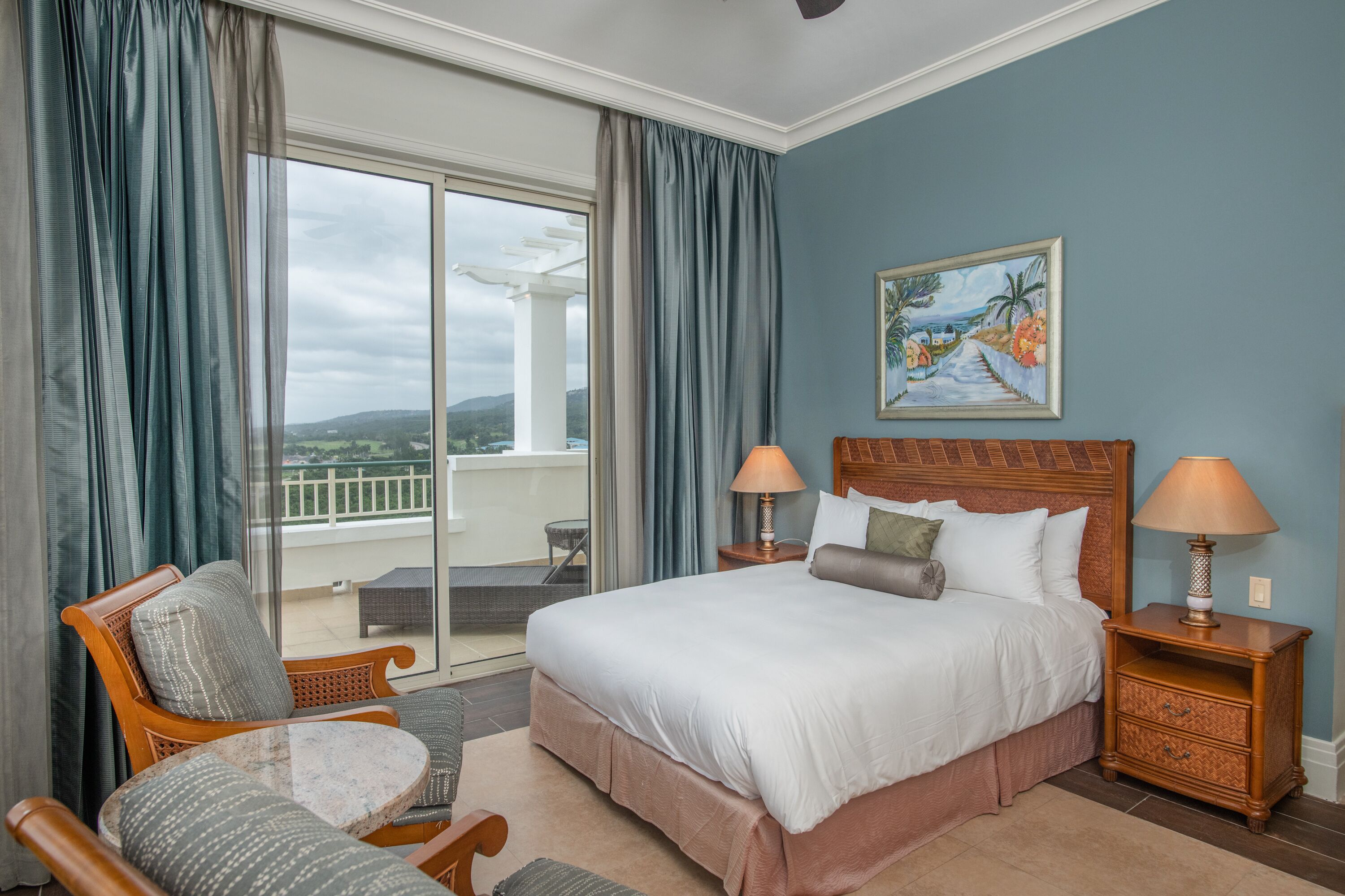 Een hotelkamer met een groot bed, twee nachtkastjes met lampen, een stoel, een balkon met uitzicht op het landschap en een schilderij aan de muur.