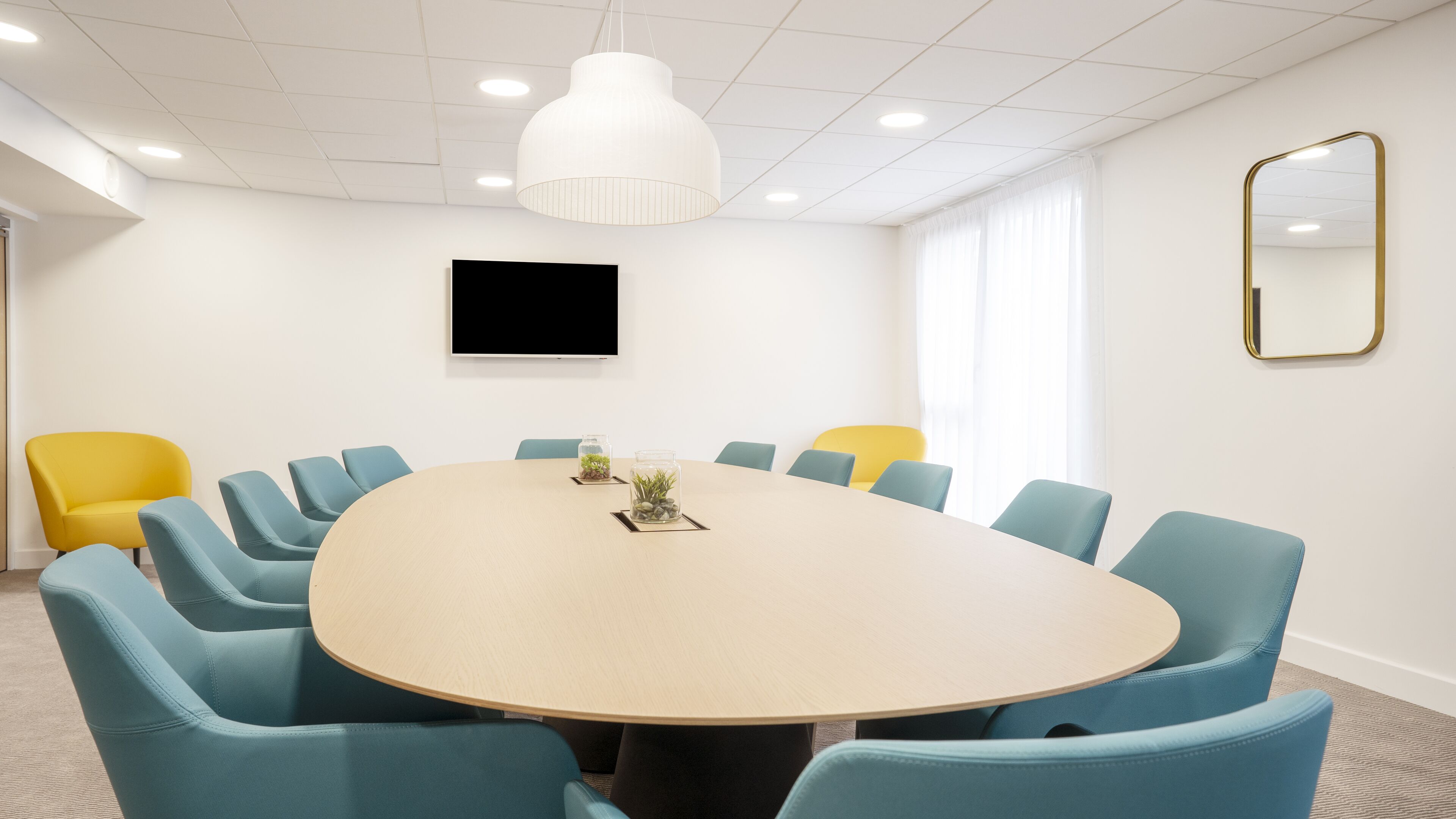 Un salón de conferencias con una mesa redonda, sillas azules y una silla amarilla, un televisor en la pared y un espejo grande.