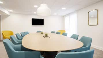 Un salón de conferencias con una mesa redonda, sillas azules y una silla amarilla, un televisor en la pared y un espejo grande.