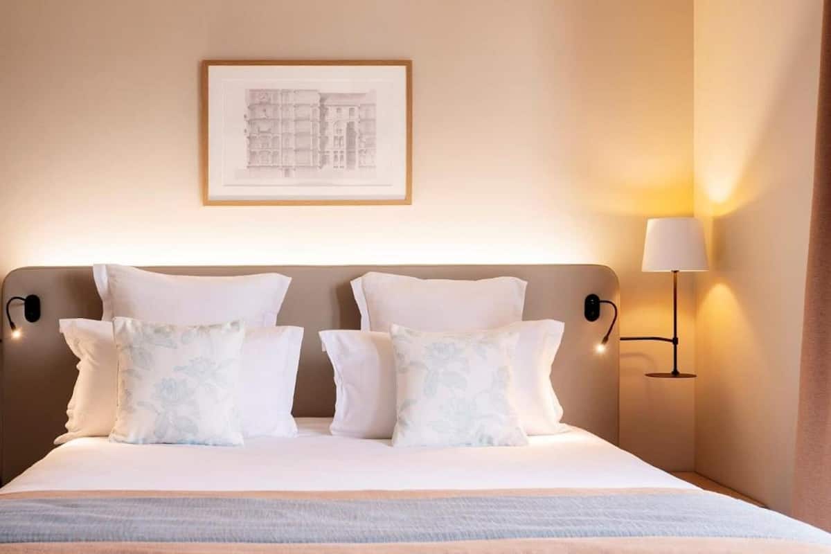 A hotel room with a bed, pillows, a framed picture on the wall, and a bedside lamp.