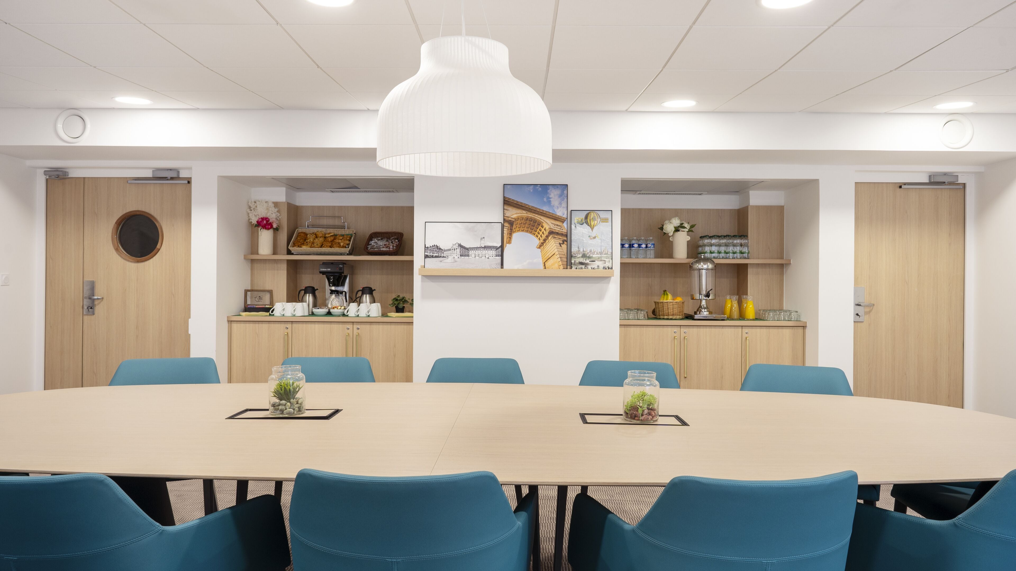 A conference room with a large oval table, blue chairs, a white pendant light, and a kitchenette with shelves above.